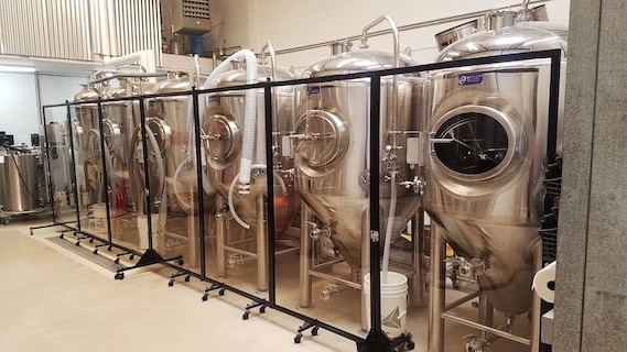 Mobile plexiglass partitions positioned in front of stainless steel brewing tanks.