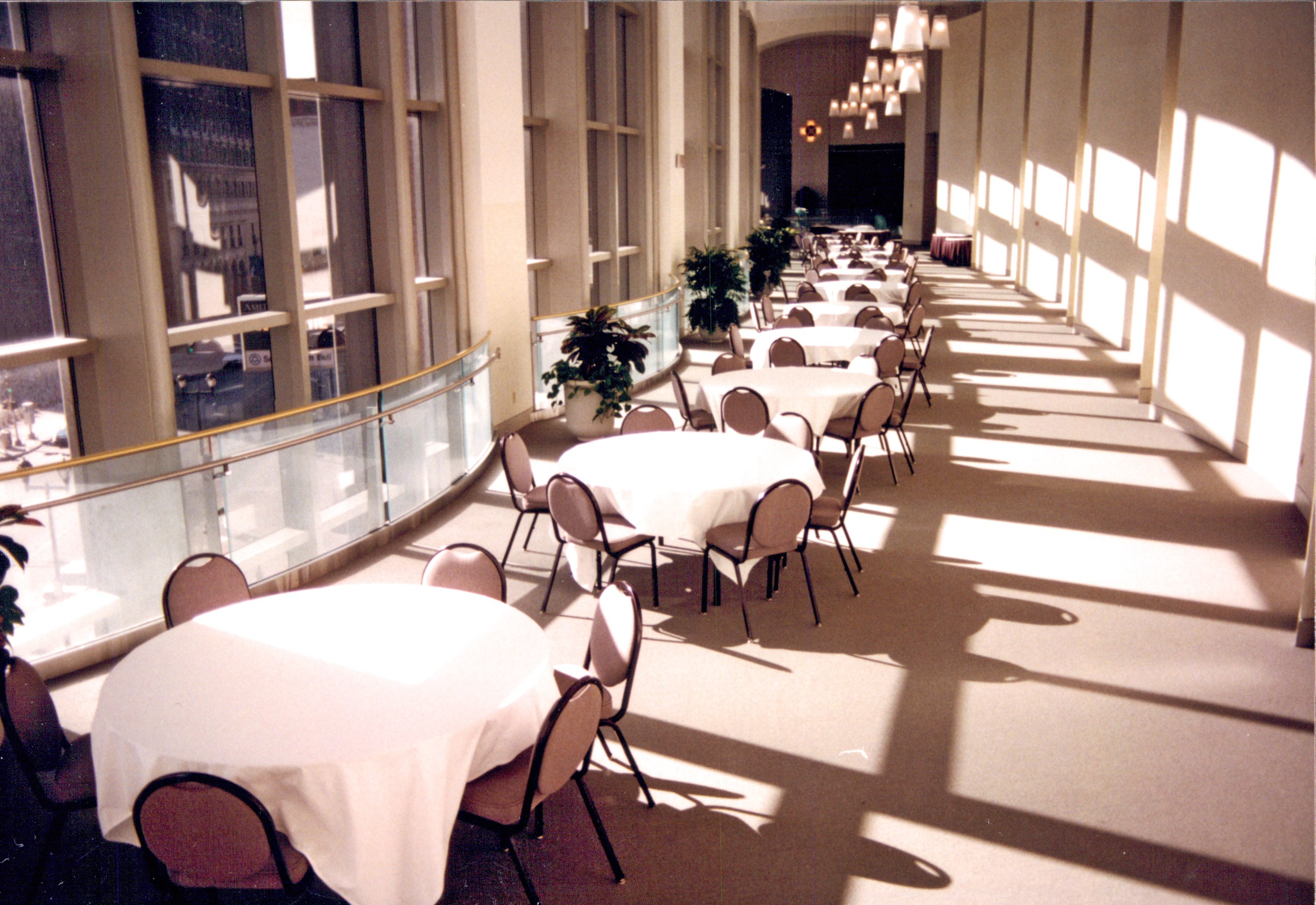 A long row of round banquet tables and chairs lined up next to floor to ceiling windows in a hallway