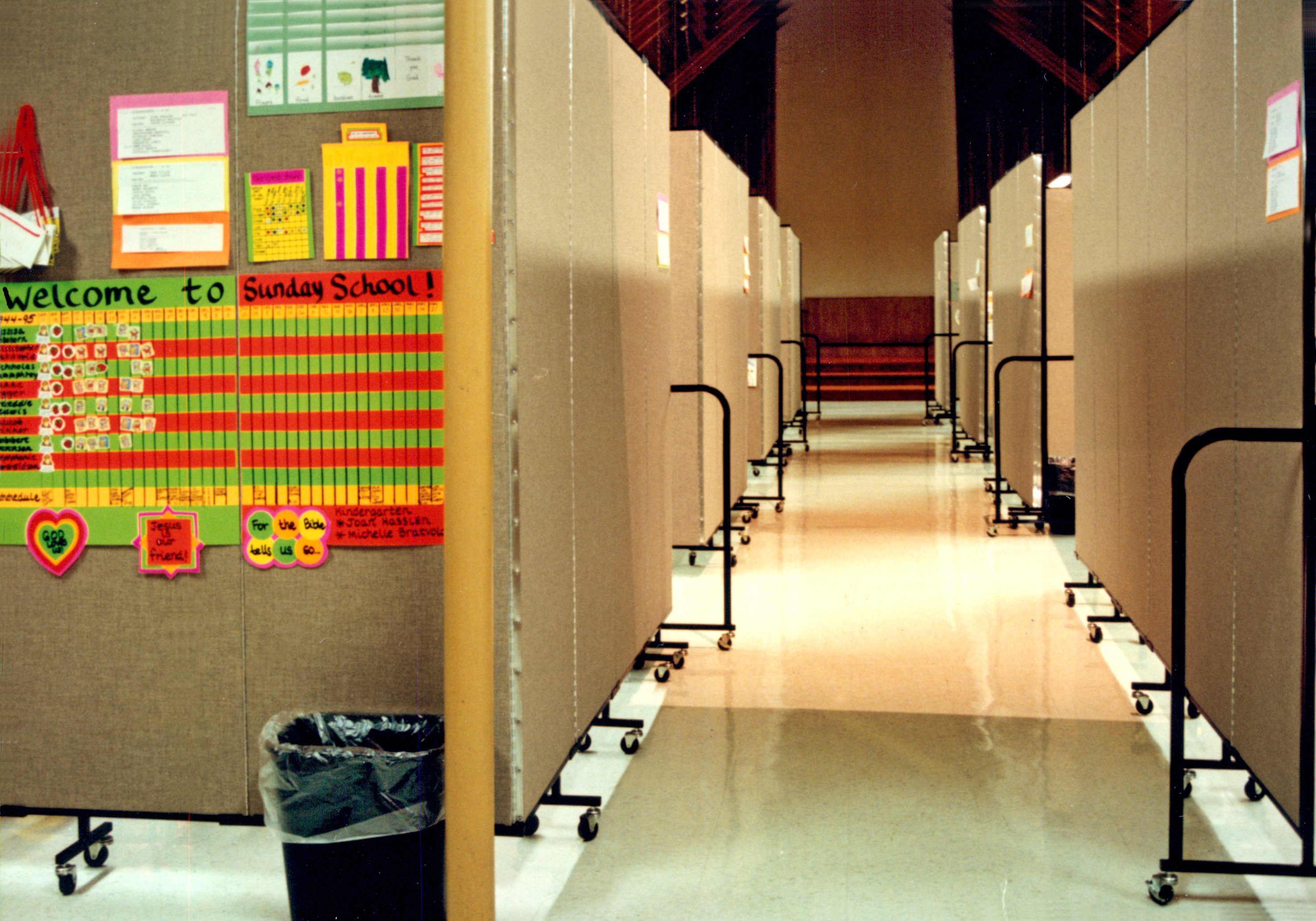 A Sunday school check in poster is hung on a room divider which is part of a row of four dividers creating Sunday School classrooms in a church