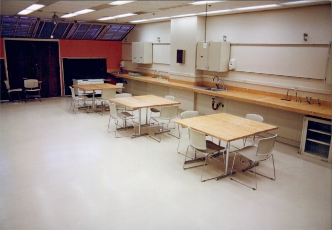A room with 3 small rectangular tables and chairs next to a counter with sinks