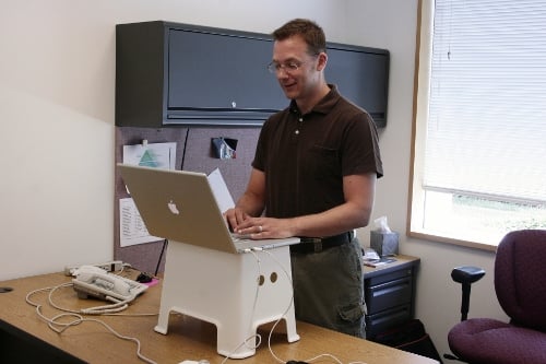 A man is standing working on his laptop computer that is resting on footstool on top of his desk