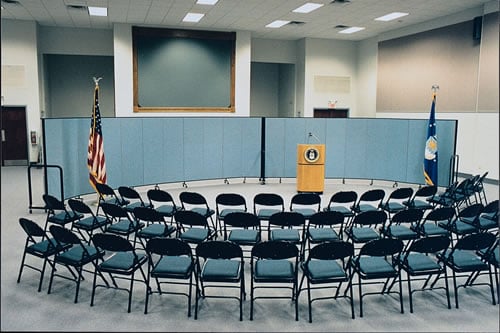 Two Screenflex Room Dividers form a curved wall in a makeshift US governement press room.