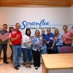 A grouping of adult men and women hold small American flags in front of a Screenflex sign