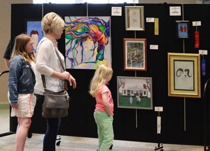 A women and two girls view a collection of artwork hung on a black portable wall