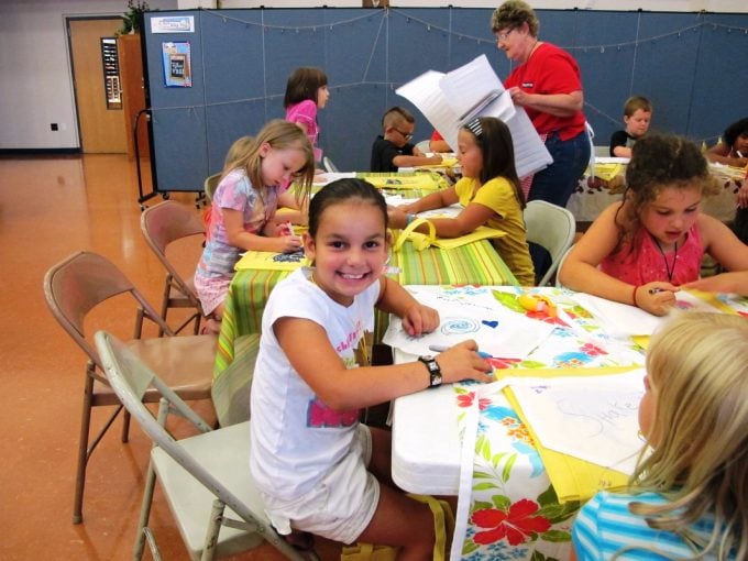 A young girl smiles at the camera as she draws a picture for her teacher