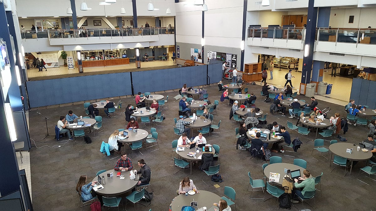 Students sit at round tables in a large room while a set of blue room dividers restrict access to other side