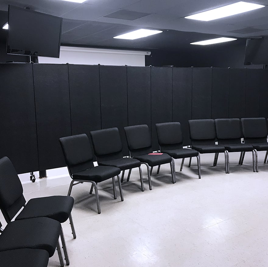 Black portable partition divider opened in a classroom with a set of chairs set in a row in front of it.