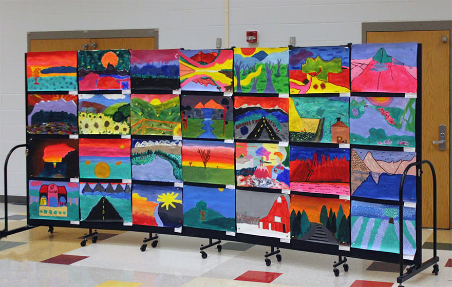 A black portable art display wall is covered with elementary school artwork.