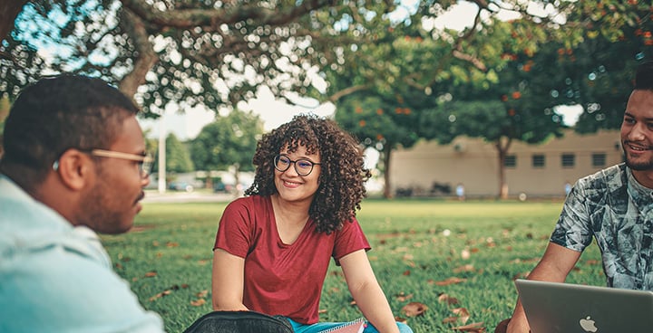 three college students smiling at each other on a lawn