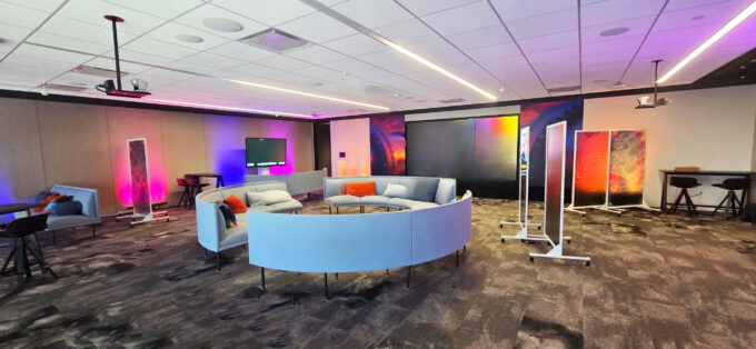 Modern lounge with circular sectional seating, colorful ambient lighting, digital displays, and high tables in a tech-enabled workspace.
