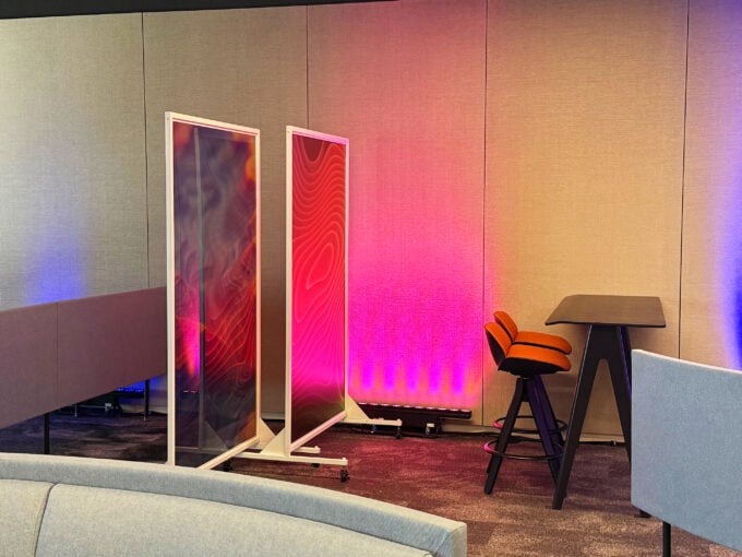 Abstract panels on wheeled frames beside a high table and stools, with colorful pink and blue lighting in a modern lounge.